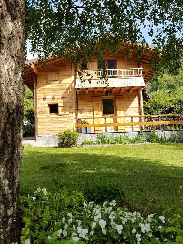 Una cabaña de madera con balcón, rodeada de un jardín cuidado y árboles.