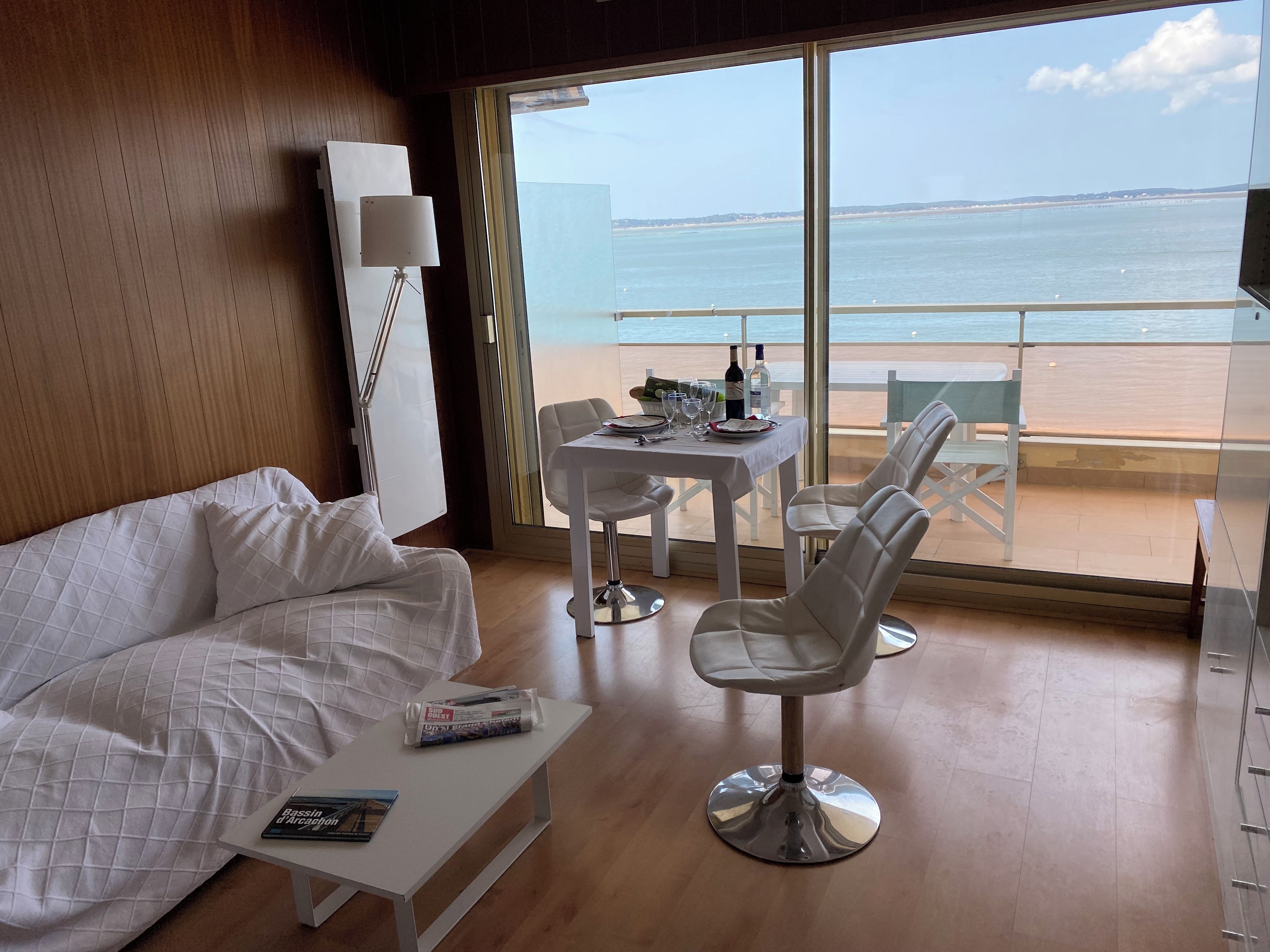 A room with a white sofa, a small table with a magazine, and a dining area with chairs and a table set for two.