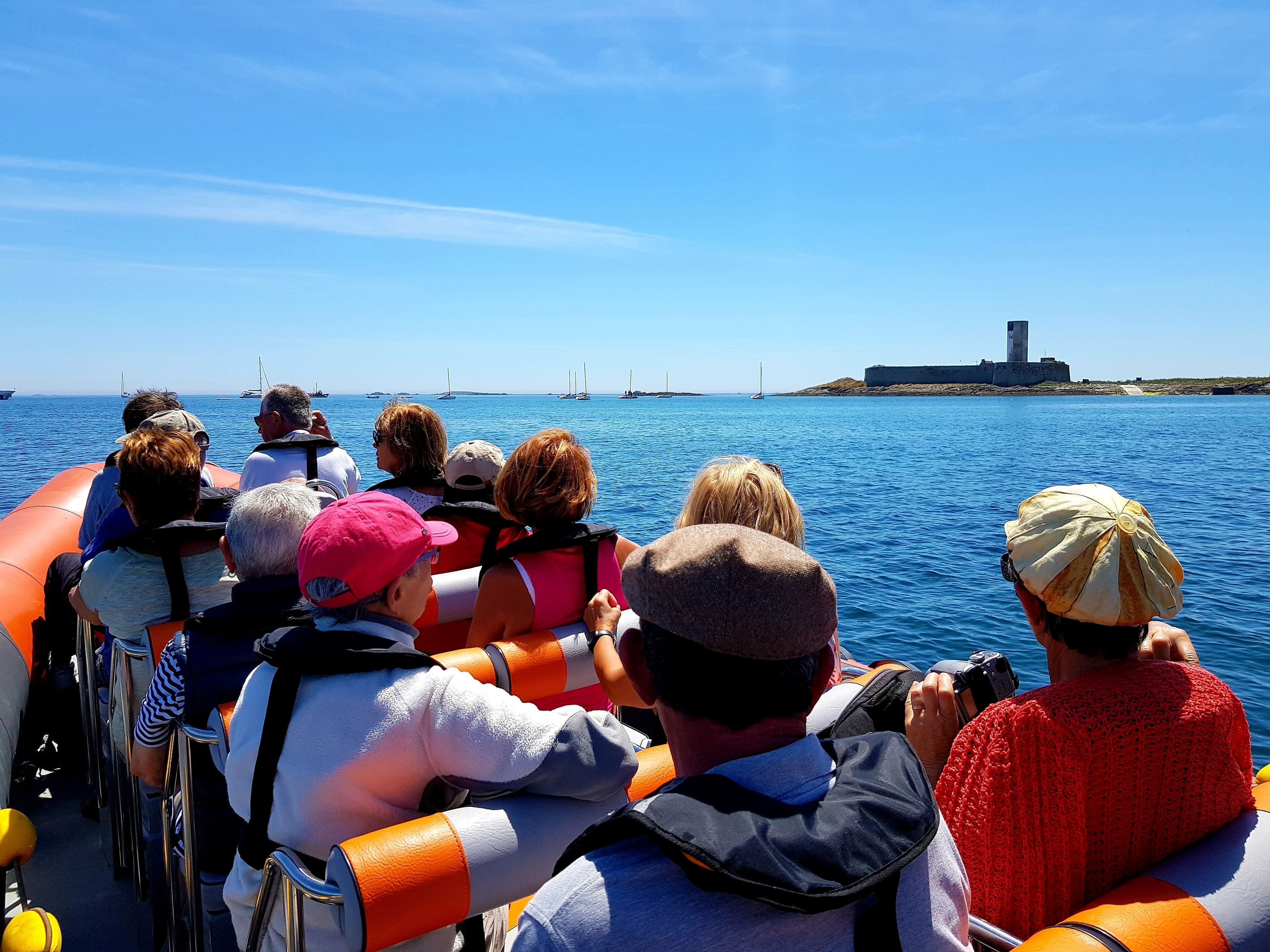 A group of people on a boat, facing a body of water with sailboats and a lighthouse in the distance.