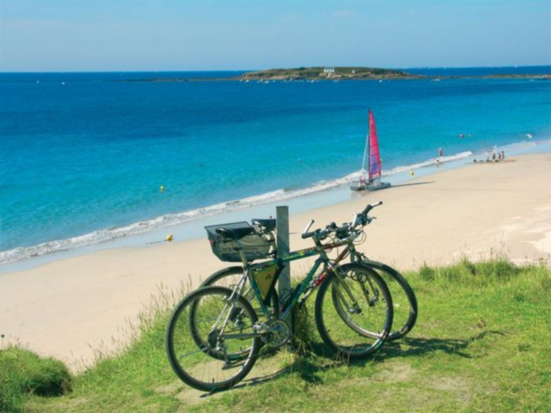 A bicycle parked on a beach with a view of the ocean and a distant sailboat.