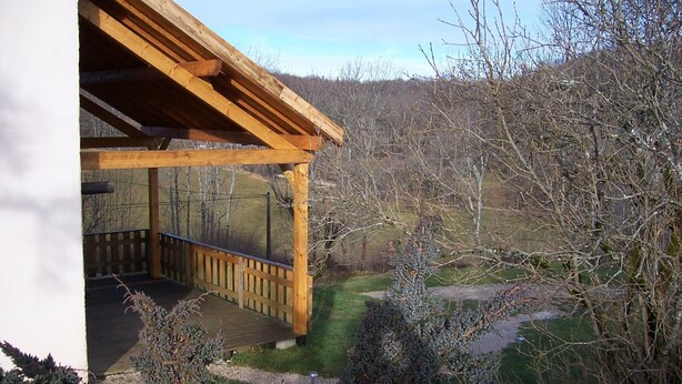Eine Holzterrasse mit Geländer, umgeben von einem Garten mit verschiedenen Sträuchern und freiem Blick auf die umliegende Landschaft.