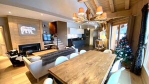 A modern living room with a wooden dining table, white chairs, a fireplace, a TV, and a Christmas tree.