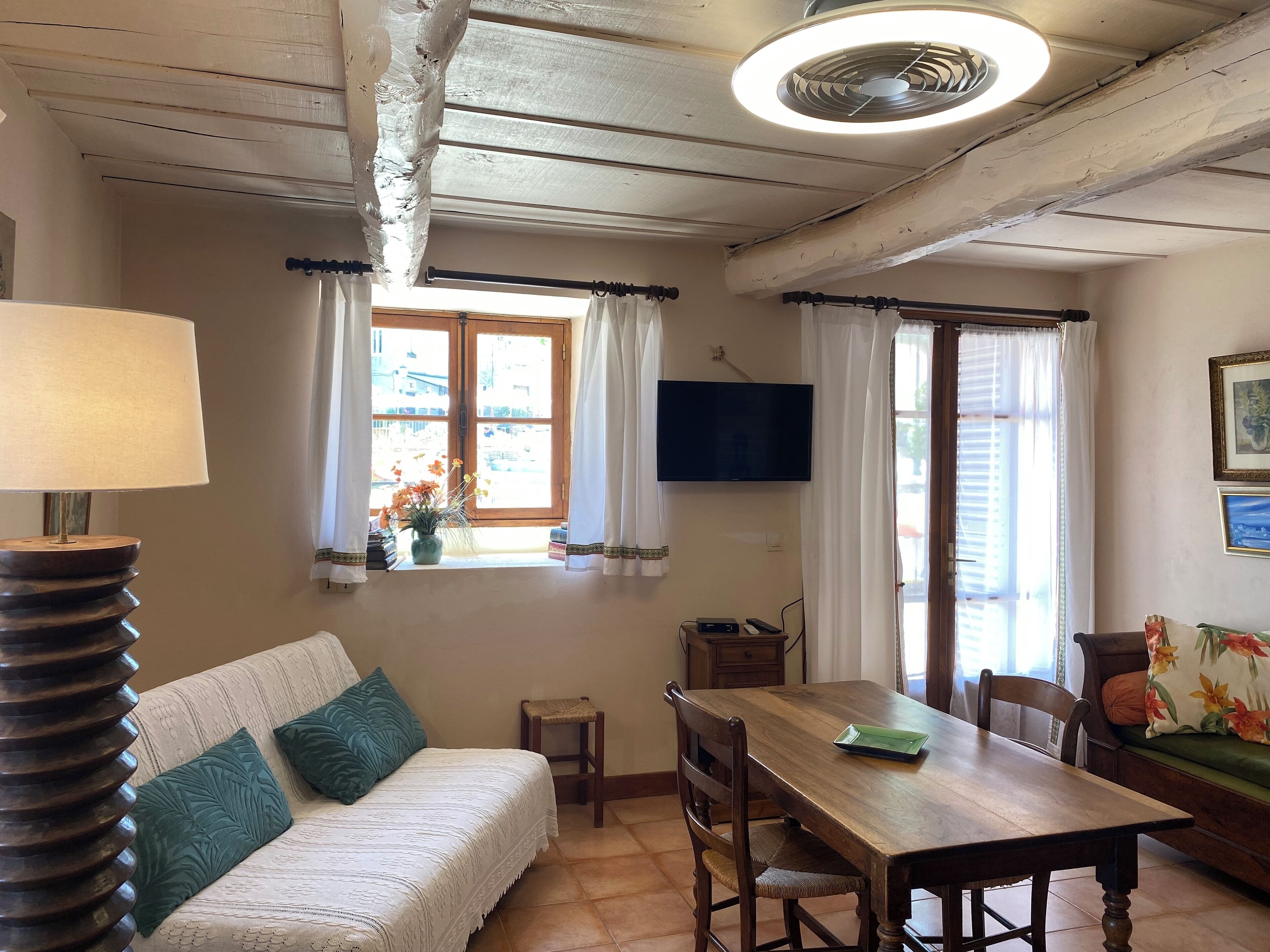 A cozy room with a wooden ceiling, a sofa with green cushions, a dining table with chairs, and a TV mounted on the wall.