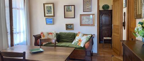 A dining area with a wooden table and chairs, a green sofa with floral cushions, and a wall adorned with framed pictures.