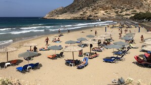 A beach with sun loungers and umbrellas, a rocky hill in the background, and the ocean. - Summer Breeze (Karpathos)