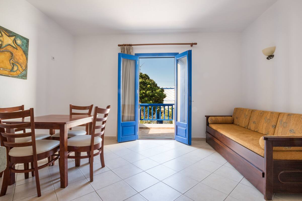 A room with a dining table and chairs, a sofa, and an open door leading to a balcony with a view of greenery.
