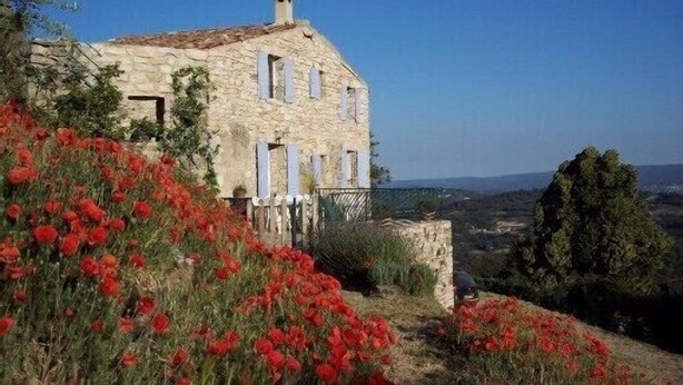 Una casa de pedra amb teulada inclinada, envoltada de pavons vermells i vista a la campanya.