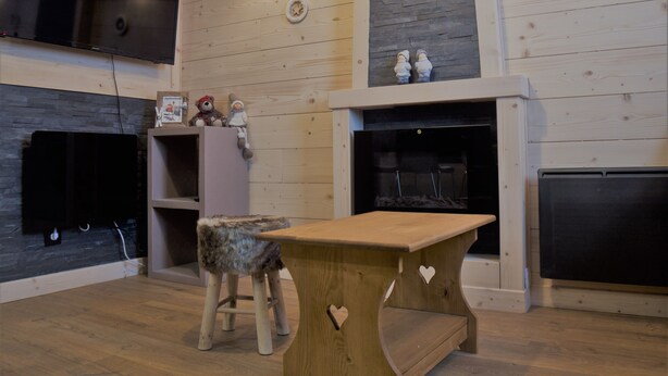 Una habitación acogedora con piso de madera, chimenea, televisor montado en la pared y una pequeña mesa con un corte en forma de corazón.