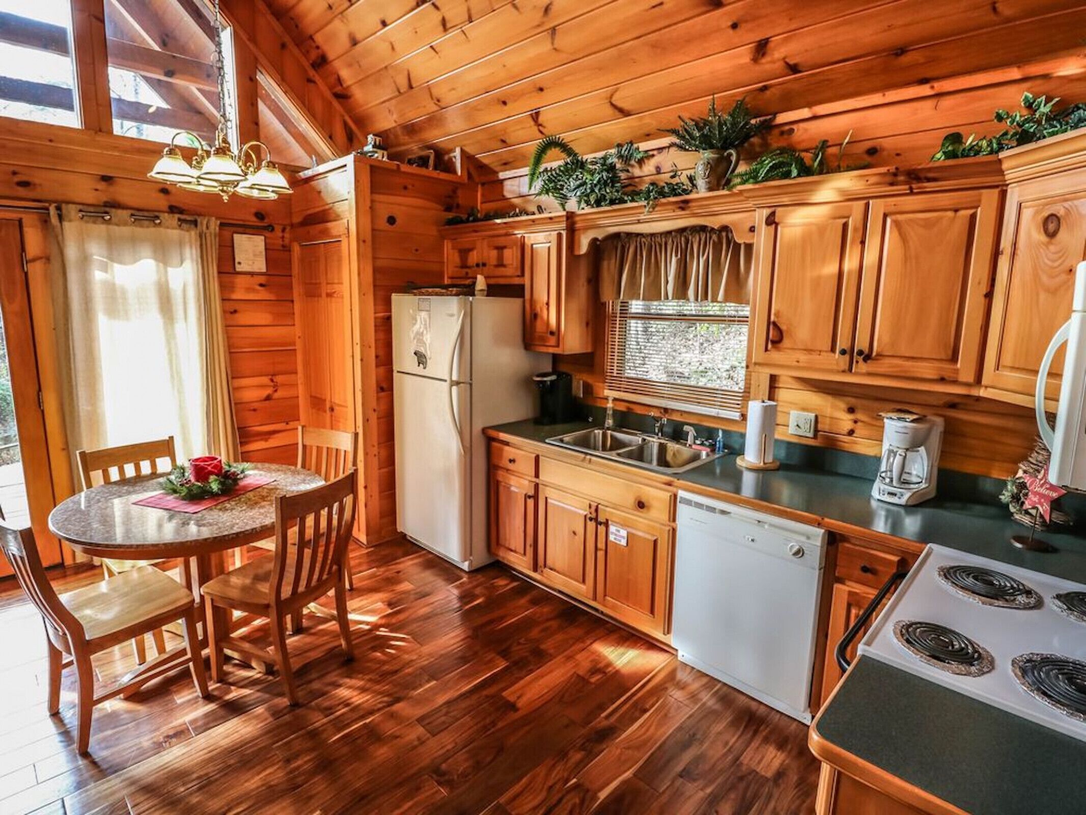 A wooden cabin kitchen with a dining table, chairs, a refrigerator, a sink, a stove, and a window with blinds.