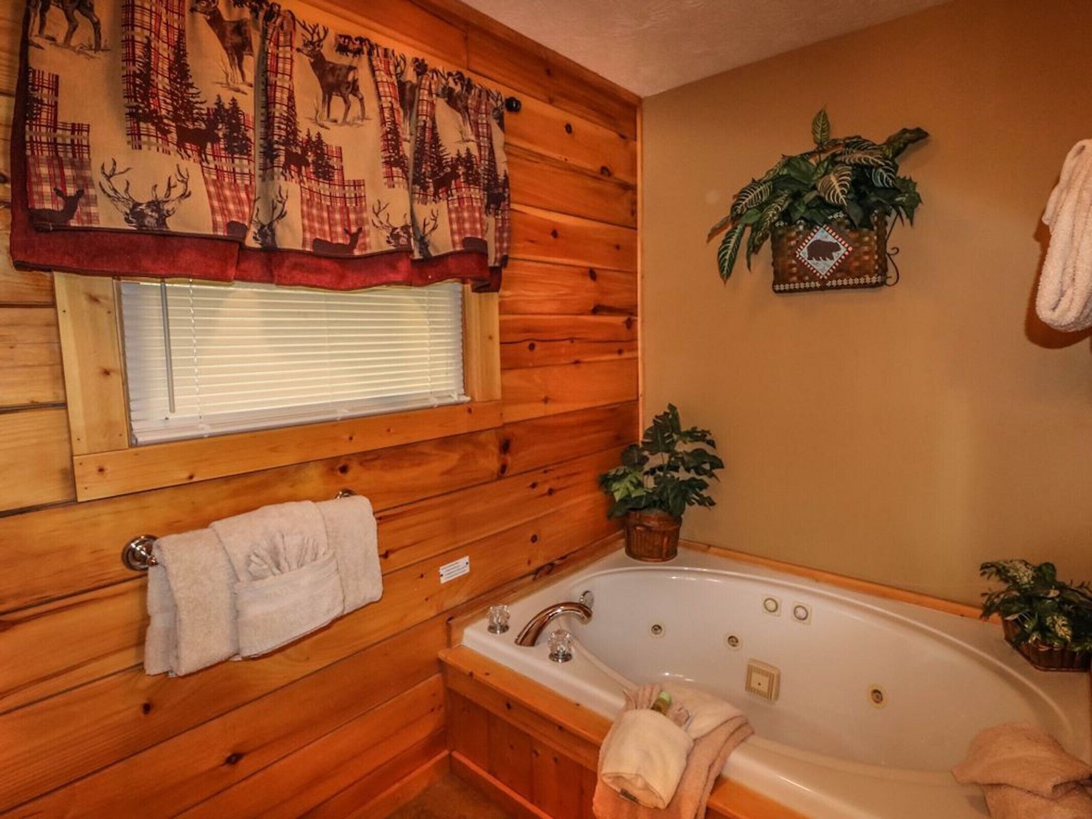 A wooden cabin bathroom with a bathtub, a window with blinds, a towel rack, and a potted plant.