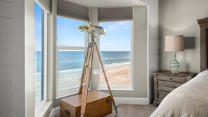 Een slaapkamer met een groot raam dat uitkijkt op een strand, een houten statief met een telescoop, een nachtkastje met een lamp, en een bed met een beddegoed met een patroon.