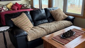 A cozy living room with a leather sofa, a wooden coffee table, and a large window with curtains. - Quiet, Cozy Lake Cottage North of Merrifield, Great Getaway! (Merrifield)