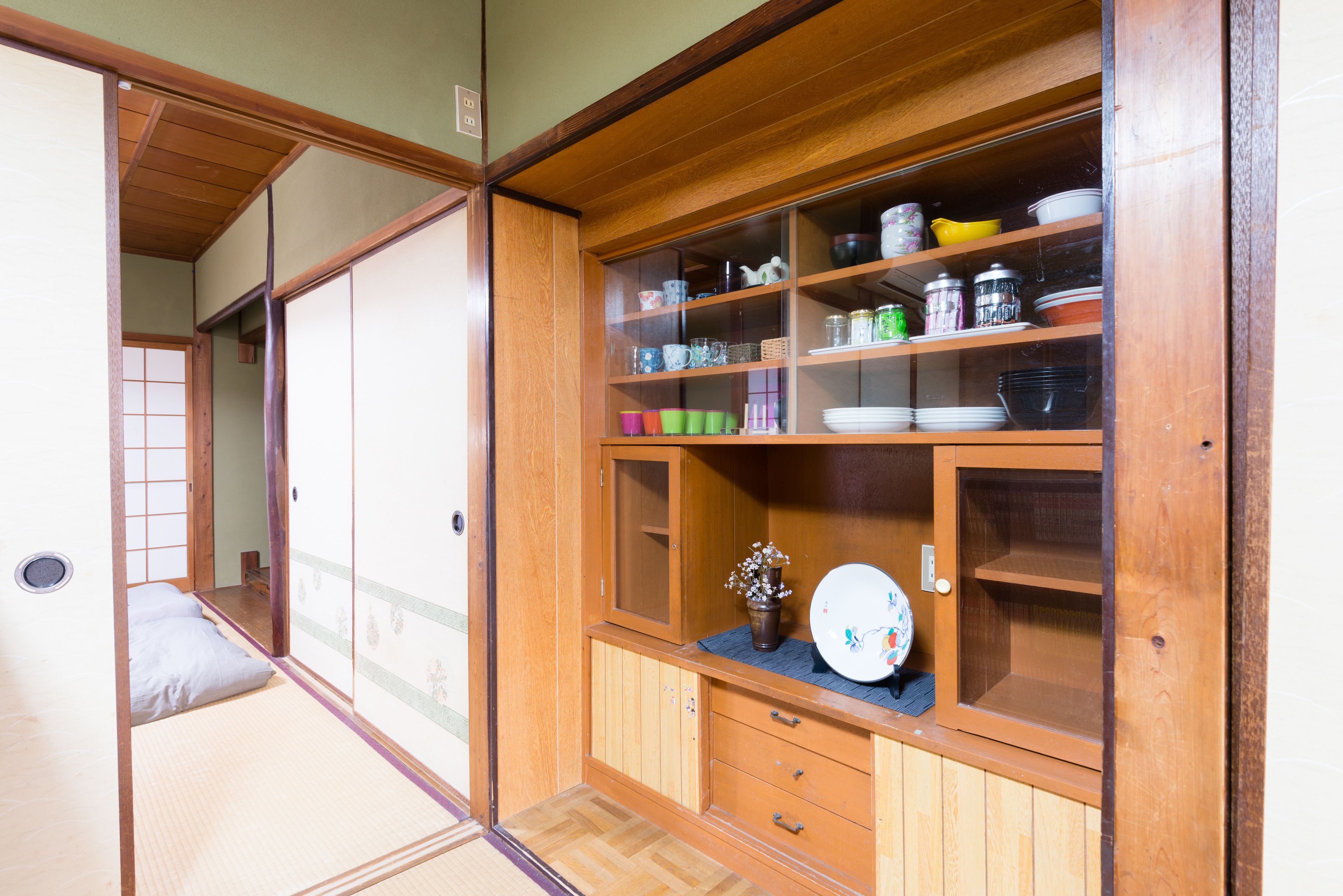A traditional Japanese room with tatami flooring, a sliding door, and a wooden cabinet with glass doors containing dishes and kitchenware.