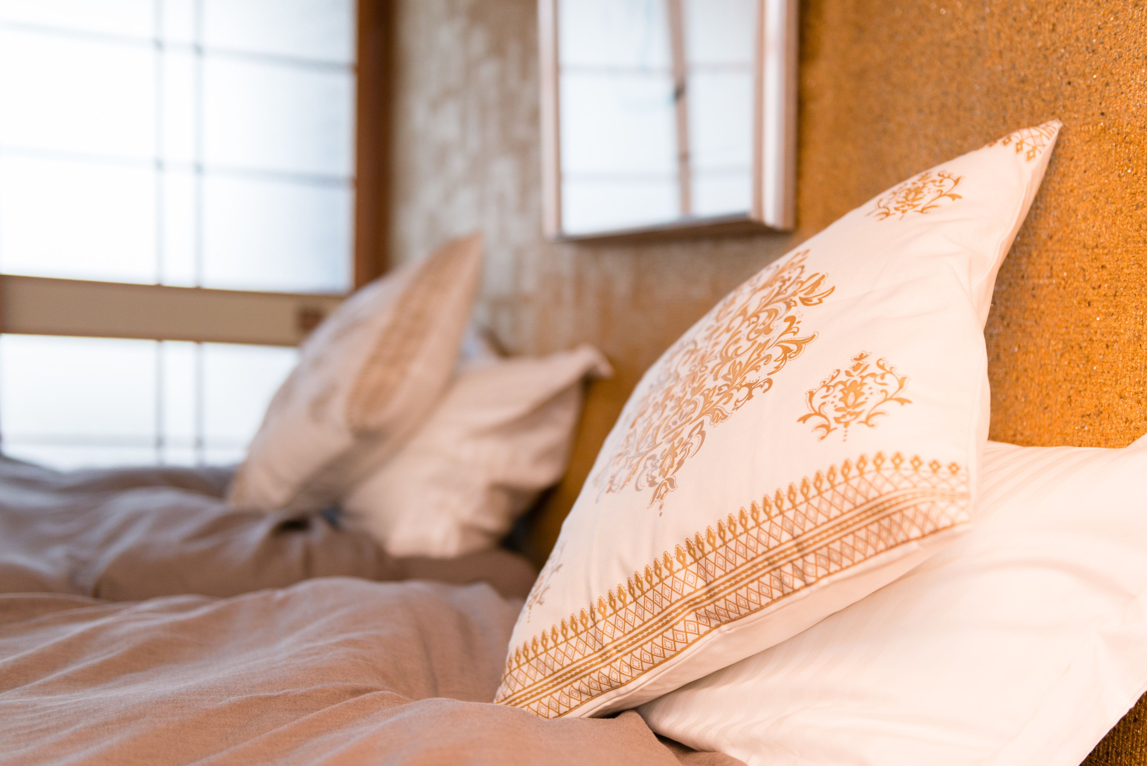 Un lit avec une literie blanche et beige, un coussin décoratif à motifs géométriques et, en arrière-plan, une fenêtre avec des stores.
