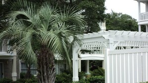 En hvid træpergola med et overdækket siddeområde, omgivet af frodig vegetation og et palmetræ.