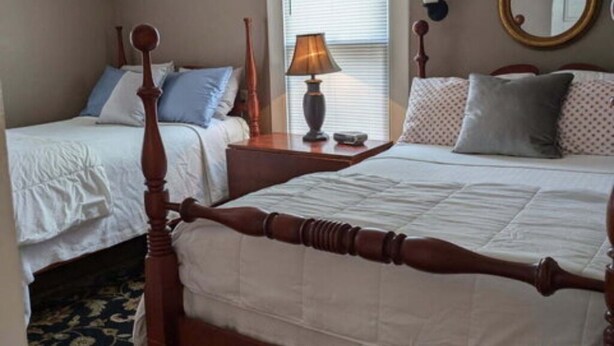 A bedroom with a large bed, a smaller bed, a nightstand with a lamp, a window with blinds, and a mirror on the wall. - Gate House (2 Doubles - 2nd Floor) (Lancaster)