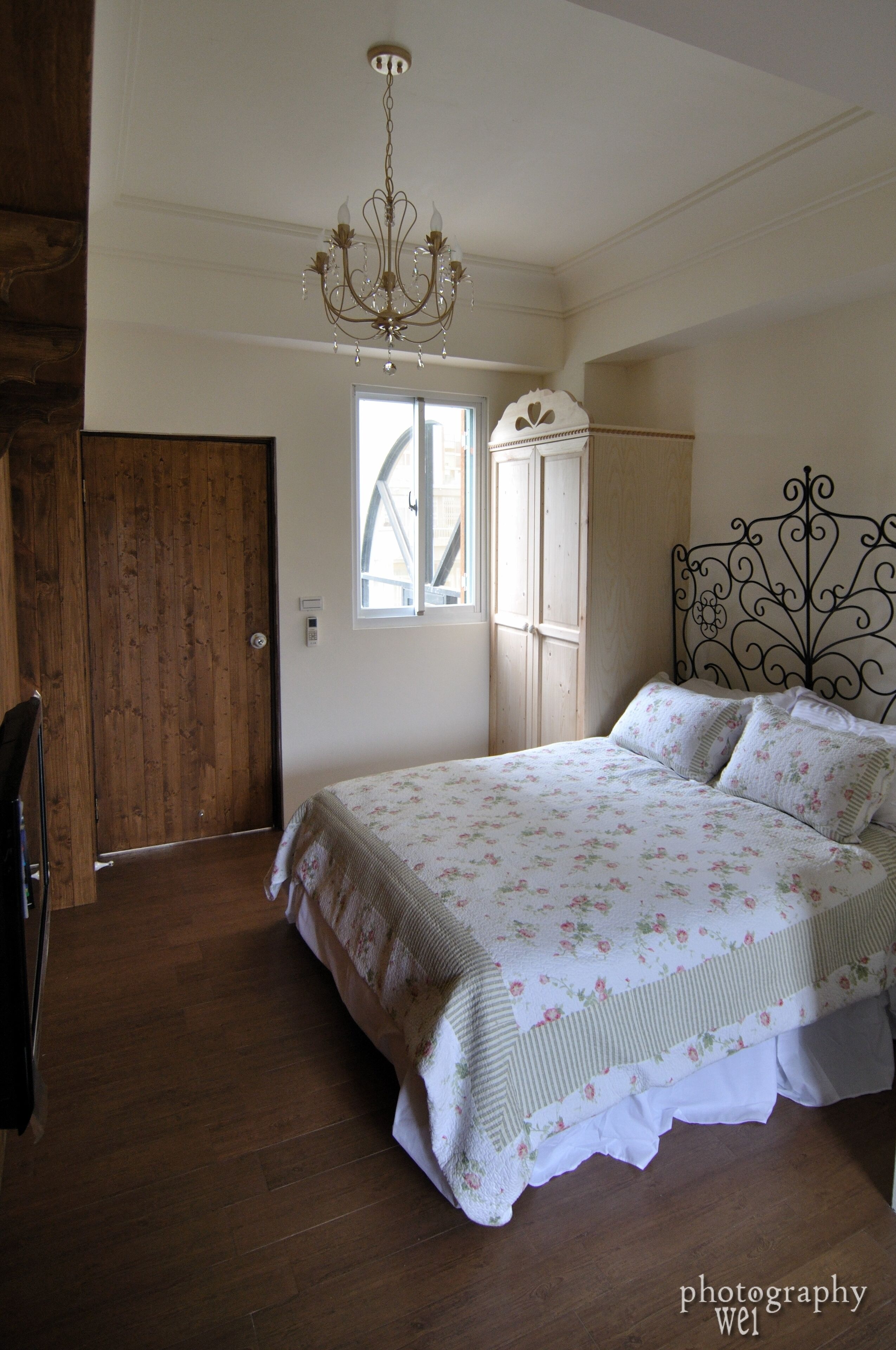 A bedroom with a wooden floor, a bed with floral bedding, a chandelier, and a window with curtains.