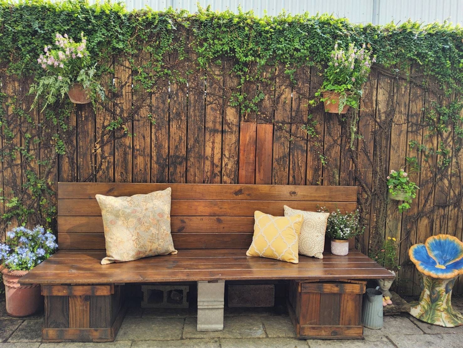 Een houten bank met kussens, tegen een houten omheining begroeid met planten.