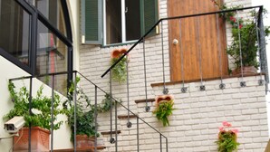 Ein mehrstöckiges Gebäude mit Ziegelverkleidung, Holztüren und grünen Fensterläden. Auf den Balkonen stehen Blumentöpfe und eine Metalltreppe ist sichtbar.