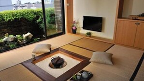 Ein Raum mit Tatami-Matten, einem Tatami-Tisch, Kissen, einem Fernseher und Blick ins Grüne.