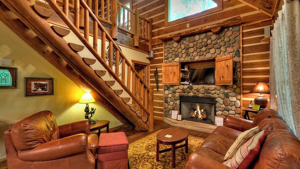 A log cabin interior with a stone fireplace, leather sofas, a wooden staircase, and a cozy ambiance. - Sans Souci | Pet Friendly (Copperhill)