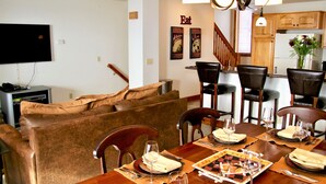 Un comedor con una mesa de madera dispuesta para cuatro personas, rodeada de sillas de cuero. Un televisor está montado en la pared, y hay una escalera que conduce a otro nivel.