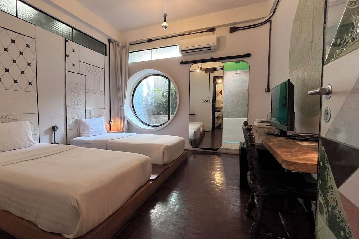 Une chambre d’hôtel avec deux lits, une fenêtre ronde, un miroir, un bureau et une télévision.