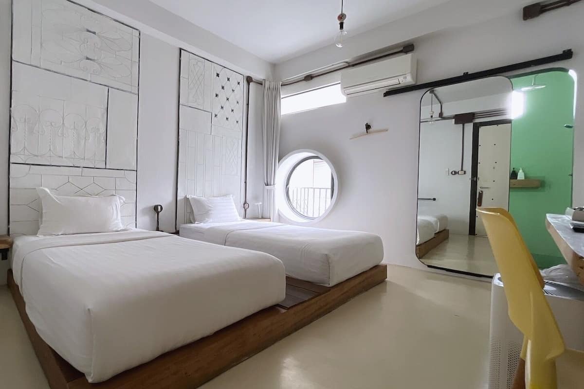 Une chambre d’hôtel moderne avec deux lits, une fenêtre ronde, un miroir et une porte verte.