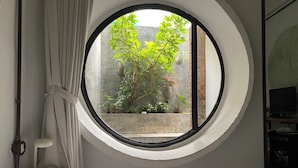 Een slaapkamer met een rond raam dat uitkijkt op een binnentuin met planten en een bakstenen muur.