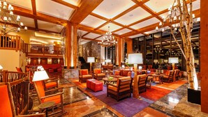 Un hall d’entrée spacieux avec un plafond haut, des poutres en bois et un lustre. On y trouve plusieurs coins salon avec des canapés et des fauteuils, ainsi qu’une cheminée en pierre.