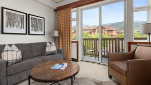 Un salon avec un canapé gris, un fauteuil en cuir marron, une table basse ronde en bois et une grande porte-fenêtre coulissante donnant sur un balcon avec vue sur les immeubles et les collines.