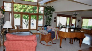 Ein geräumiges Wohnzimmer mit einem großen Holzpiano, Korbmöbeln und großen Fenstern mit Blick auf Bäume.