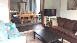 Una sala de estar con un sofá marrón, una mesa de centro y un televisor sobre un mueble bajo. Se observa un comedor con una mesa y sillas al fondo.