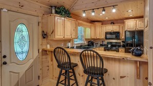 A wooden cabin interior with a kitchen featuring wooden cabinets, a bar area with two stools, a microwave, a stove, and a refrigerator. - Country Dreams | Near Ocoee River | Hot Tub | Fire Pit (Copperhill)