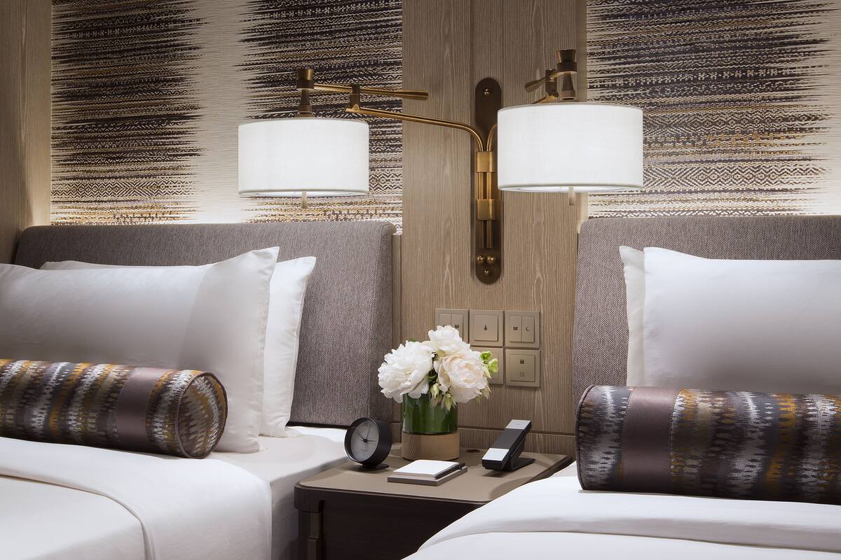 A hotel room with two beds, a bedside table with a clock, a vase with flowers, and a wall with a textured pattern.
