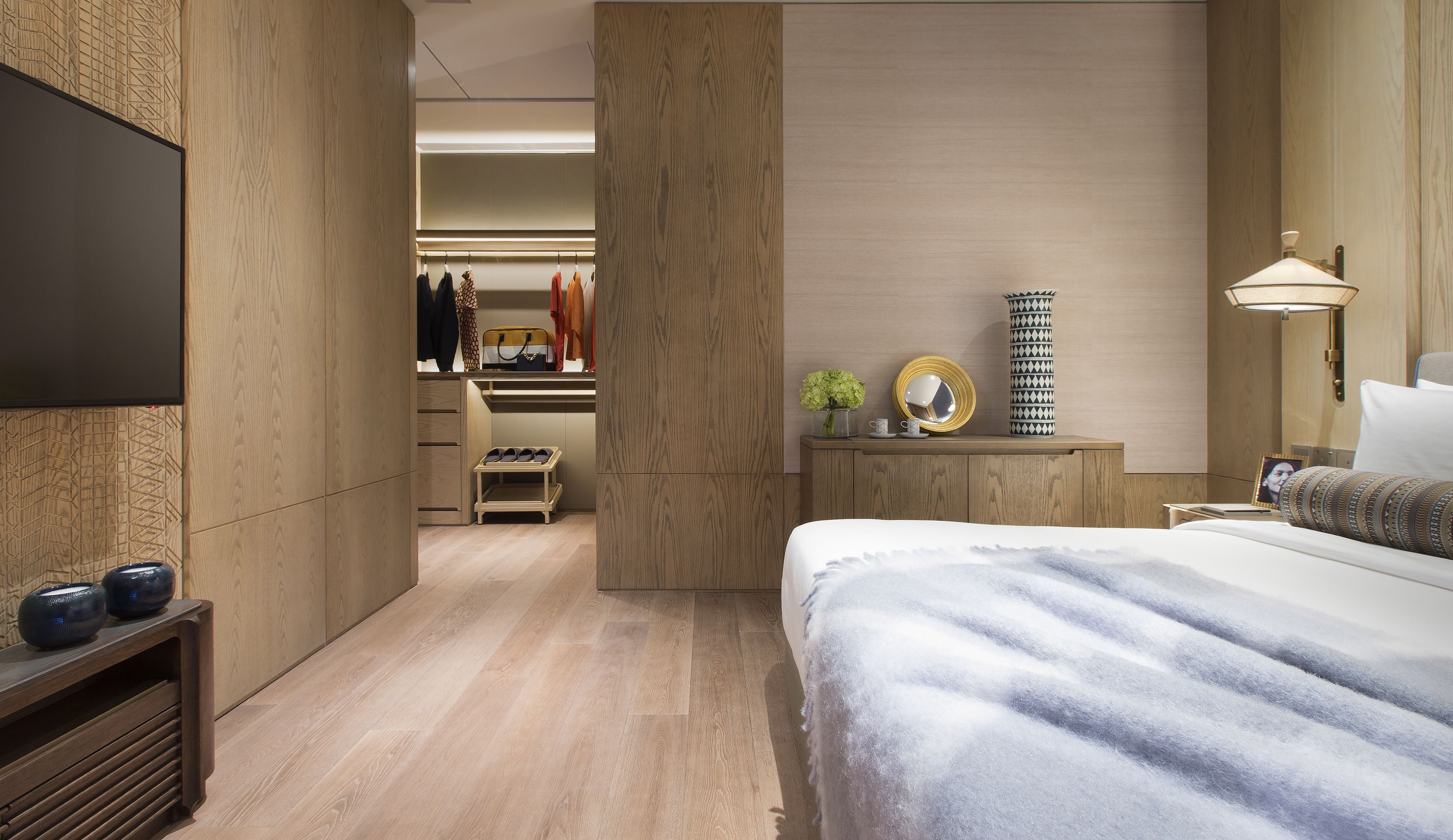 A modern hotel room with a large bed, a TV mounted on the wall, a walk-in closet, and a bedside table with a lamp.