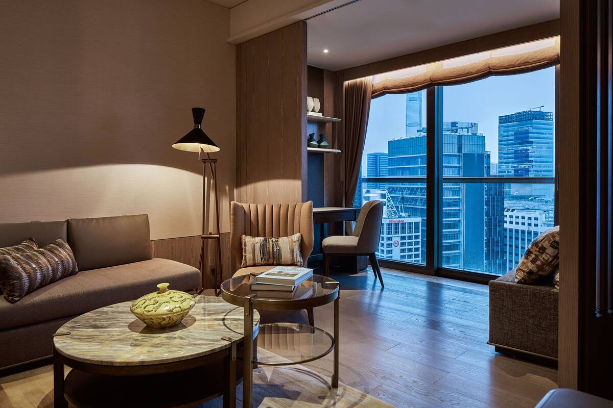 A modern living room with a sofa, armchair, and round coffee table. A large window offers a city view.