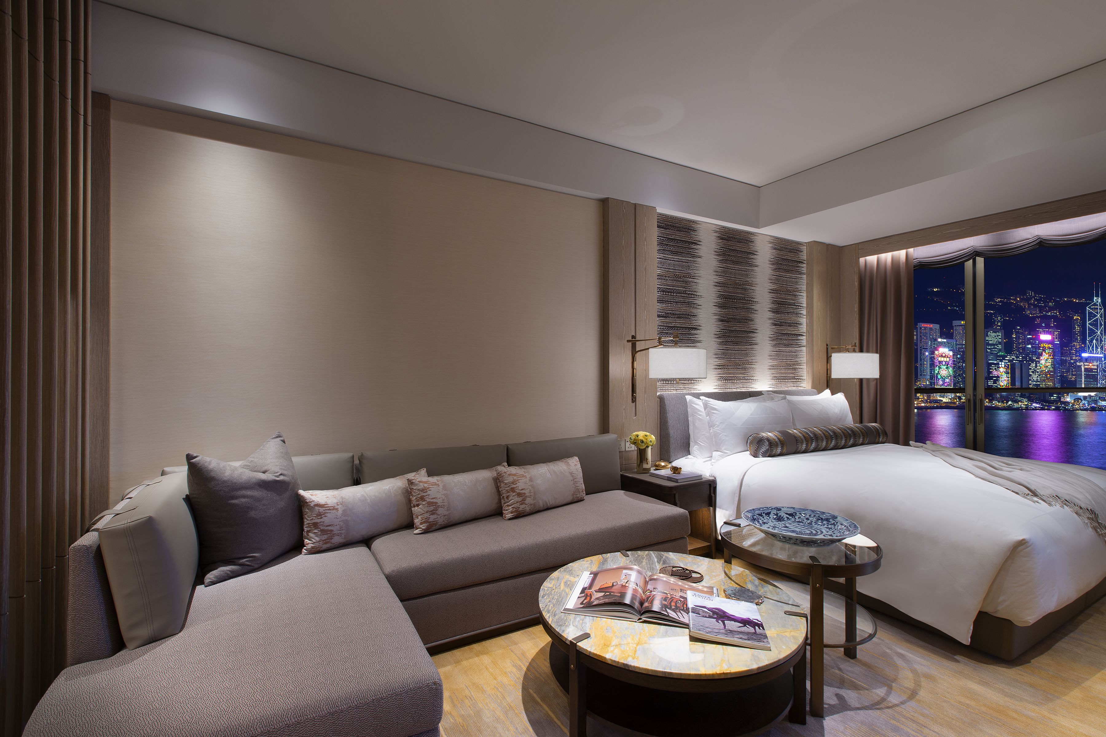 A modern hotel room with a sofa, a bed, a nightstand, and a view of the cityscape at night.