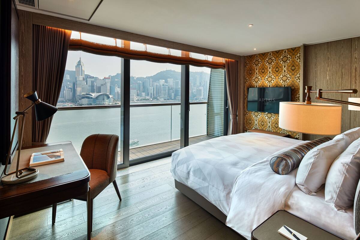 A hotel room with a large bed, a desk with a lamp, a chair, and a TV.