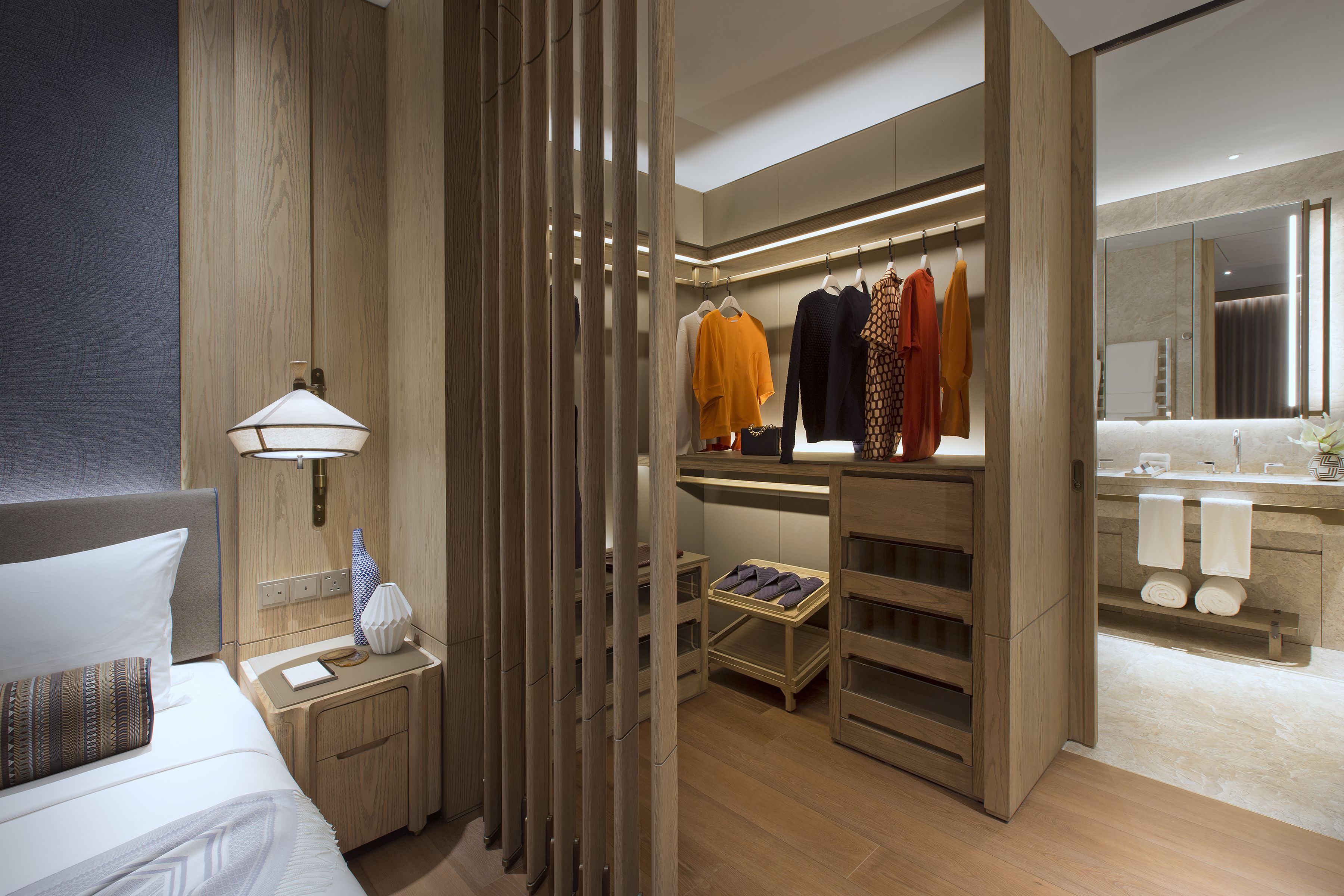 Une chambre d’hôtel moderne dotée d’une tête de lit en bois, d’une table de chevet avec une lampe, d’une armoire où sont suspendus des vêtements, et d’une salle de bain avec un miroir et un porte-serviettes.