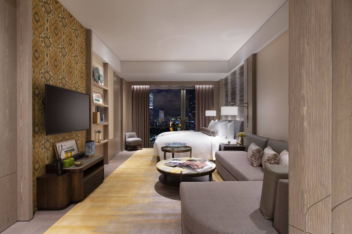 A hotel room with a bed, a TV, a sofa, a coffee table, and a view of the cityscape.