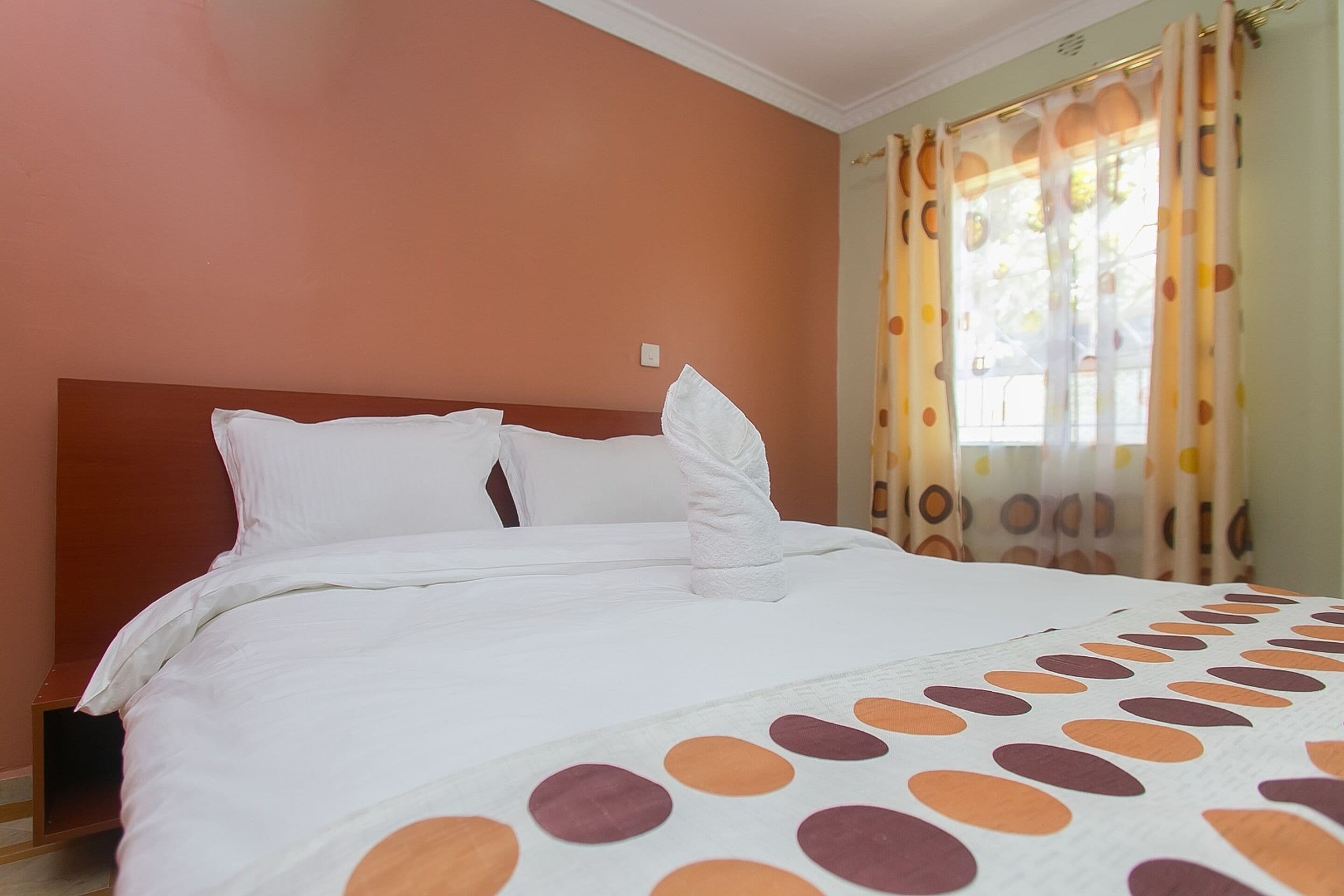 A neatly made bed with white linens and a polka dot blanket, a wooden headboard, and a window with curtains.