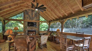 A rustic cabin interior with a stone fireplace, wooden furniture, and large windows overlooking a forested area.