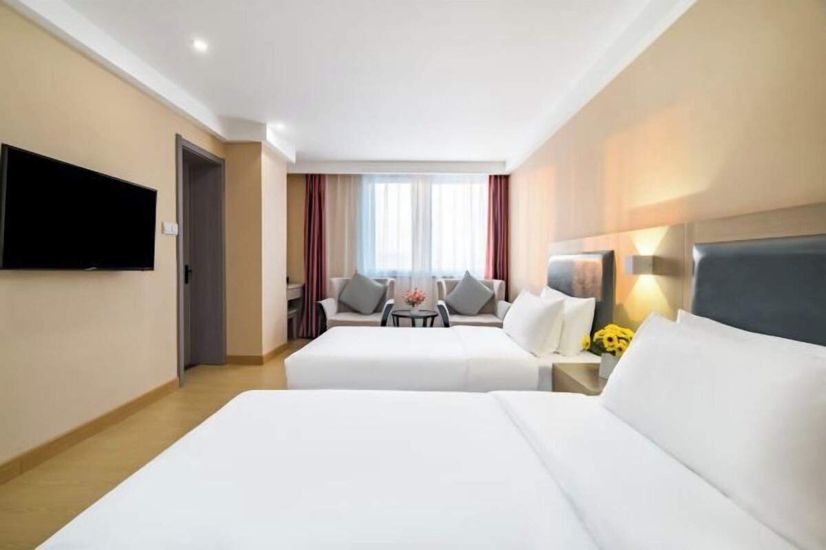 Ein Hotelzimmer mit zwei Betten, einem Fernseher, einem Sofa, einem kleinen Tisch mit Blumen und einem Fenster mit Vorhängen.