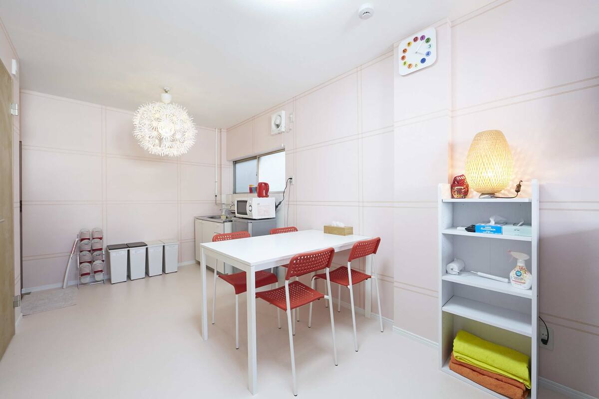 A modern kitchen with a white table, red chairs, a hanging light fixture, and a shelf with cleaning supplies.