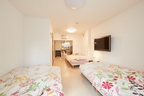 A bedroom with two beds, a TV, and a kitchenette in the background.