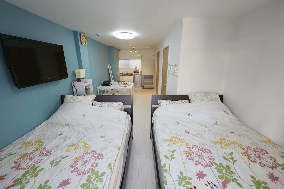 A hotel room with two beds, a TV, a desk, and a kitchenette.