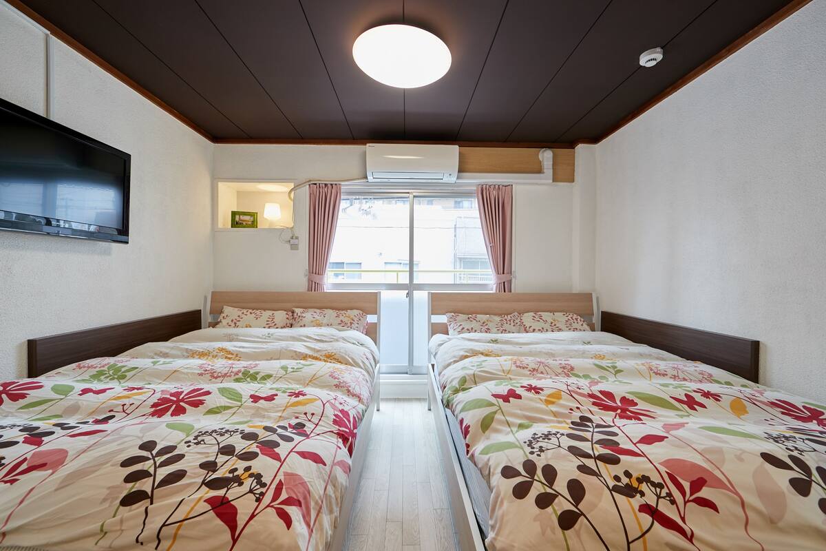 A room with two beds, a TV mounted on the wall, an air conditioner, and a window with curtains.