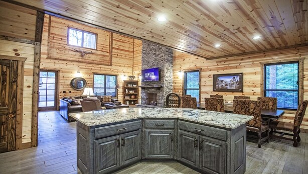 A spacious log cabin interior with a large kitchen island, stone fireplace, and a dining area with patterned chairs. - Cowboy Castle - Ridge Line Lodge, Hot Tub, Games, Outdoor Entertaining🔥 🌲 (Broken Bow)