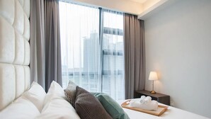 Ett hotellrum med en stor säng, grå gardiner, ett sängbord med en lampa och utsikt över byggnader genom fönstret.
